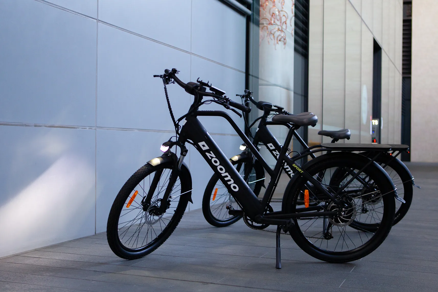 White electric bicycle with black tires and the brand name Zoomo on the frame against a black and white geometric wall background.